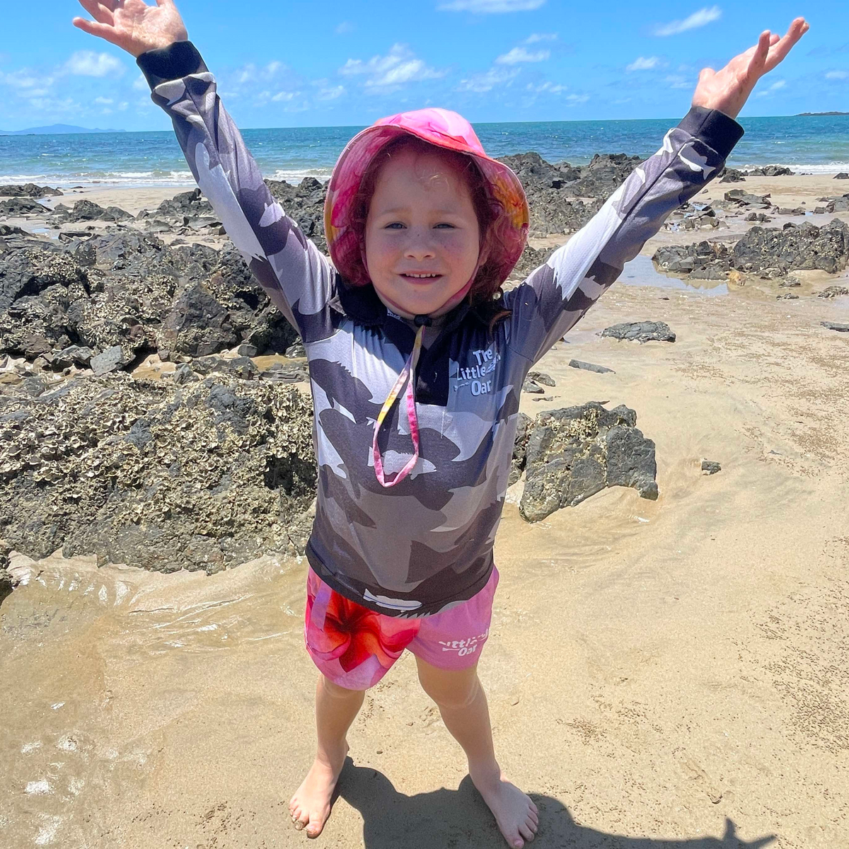 A child standing on a beach wearing The Dirty Oar Coral Trout Kids Fishing Shirt, Frangipani Kids shorts and Frangipani bucket hat, and posing with arms raised.