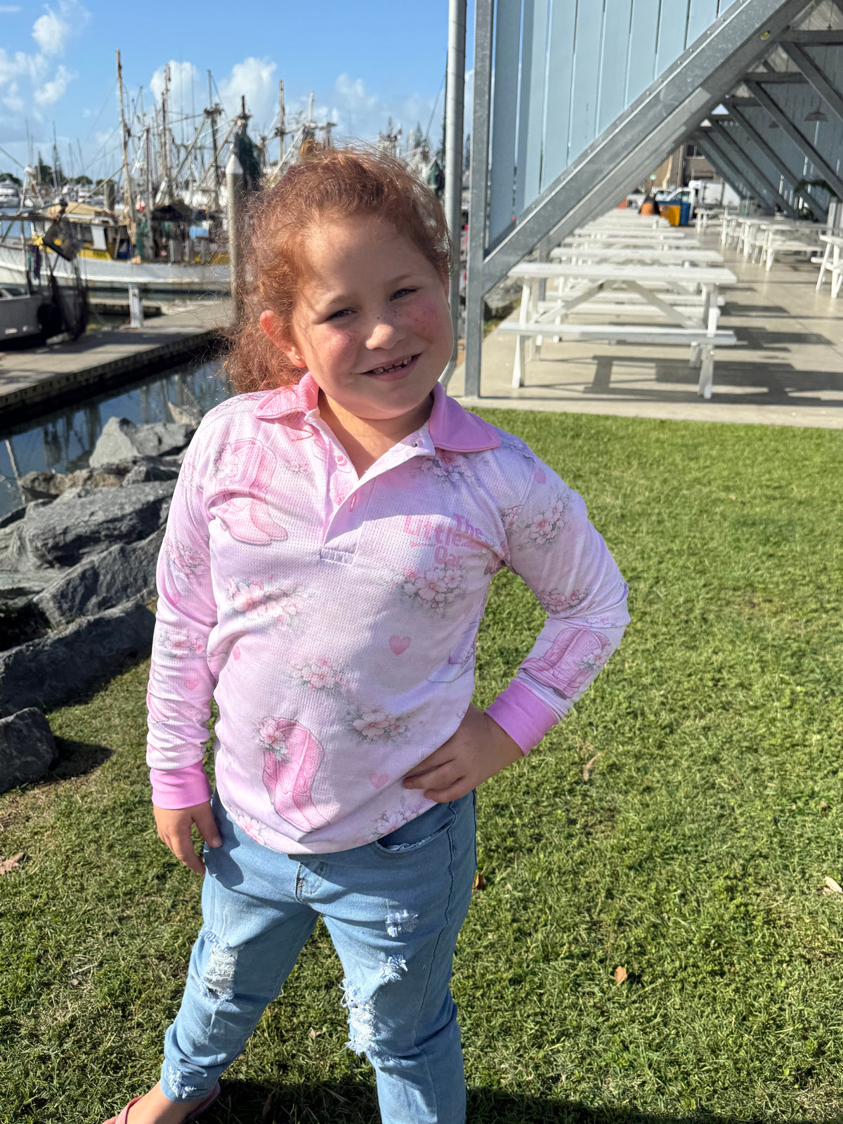 Young girl wearing The Dirty Oar Country Dreams Kids Fishing Shirt and blue jeans standing on grass near a marina.