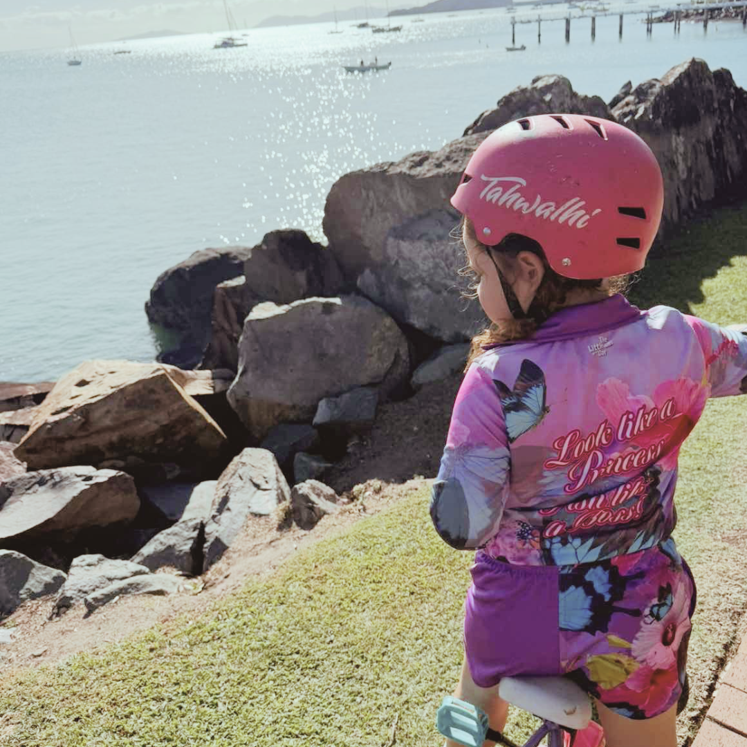 Child wearing Princess Kids Shorts  with Princess Kids Fishing Shirt and pink helmet by a waterfront with rocks and grass.