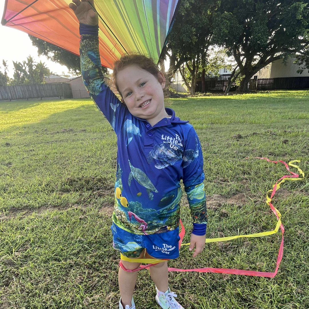 Child holding a colorful kite in a grassy outdoor setting, wearing The Reef Kids Fishing Shirt and The Reef Kids Shorts by The Dirty Oar.
