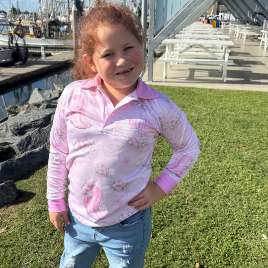 Young girl wearing The Dirty Oar Country Dreams Kids Fishing Shirt and blue jeans standing on grass near a marina.