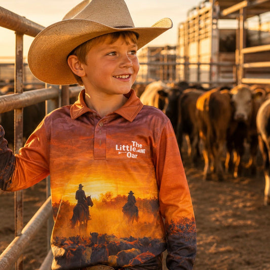Young boy in a cowboy hat wearing The Dirty Oar's Country Livin Kids Fishing Shirt with a sunset design, standing in a pen with cattle.