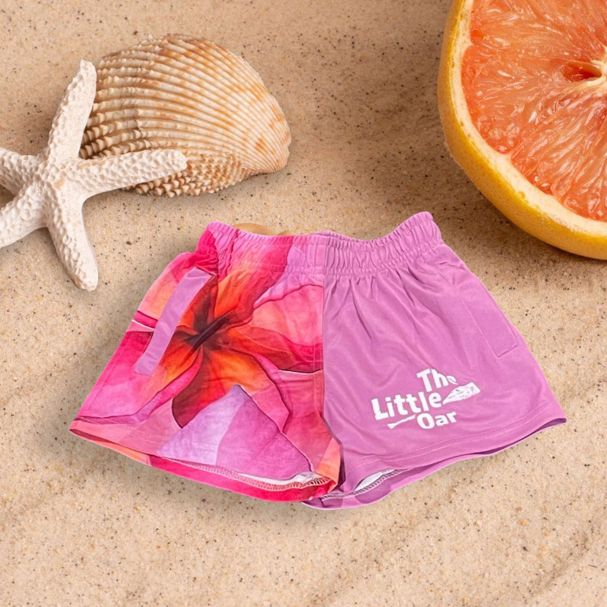 The image features a pair of The Diry Oars Frangipani Kids Shorts, laid out on a sandy surface with seashells and a slice of pink lemon nearby. The shorts are designed with a comfortable elastic waistband and deep pockets.