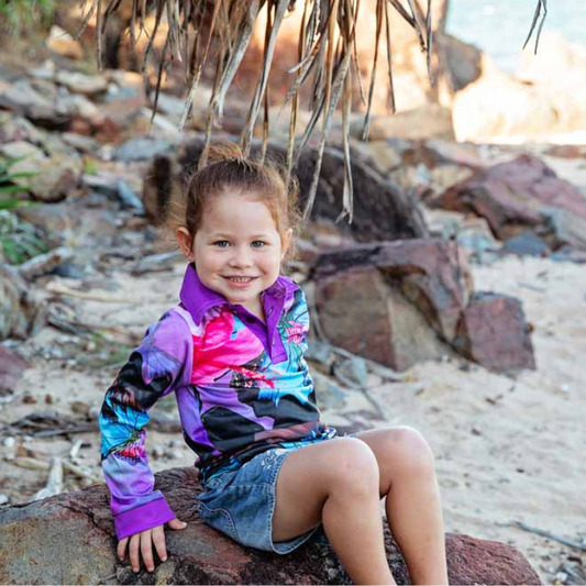 A child sitting on a rock wearing The Dirty Oars Princess Kids Fishing Shirt with a butterfly pattern and short pants, smiling at the camera.