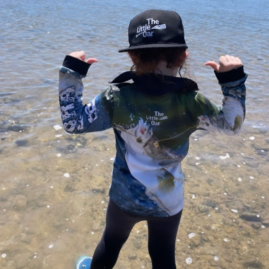 child wearing The Dirty Oar Barra Kids Fishing Shirt and The Little Oar Flat Bream cap, standing by the sea.