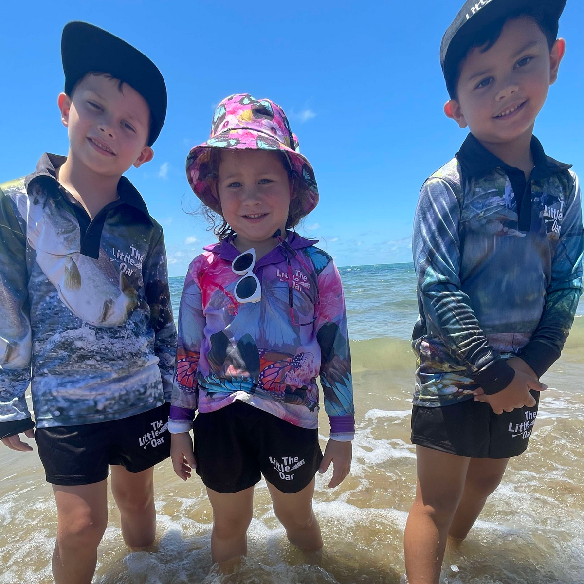 Three children standing in the water, wearing The Dirty Oar Kids Fishing Shirts and matching The Dirty Oar Kids Shorts, with sunglasses and hats on a sunny day.