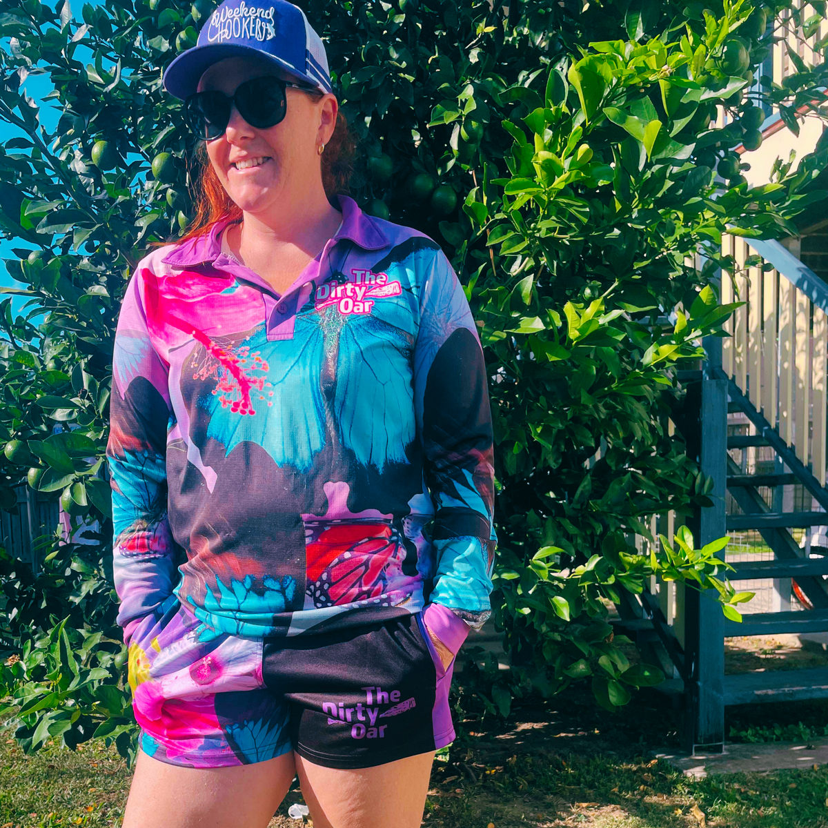 A lady wearing The Dirty Oars Princess Womens Fishing Shirt paired with matching Princess Womens shorts, standing outdoors with plants in the background.