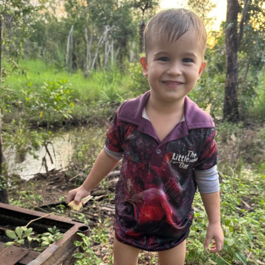 A child wearing the dirty Oar Mangrove Jack Kids Fishing Shirt, standing near water and trees.