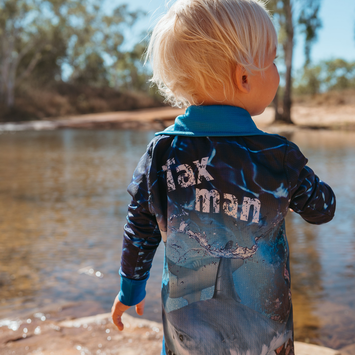 Child wearing The Dirty Oar Taxman Kids Fishing Shirt by a body of water with trees in the background.