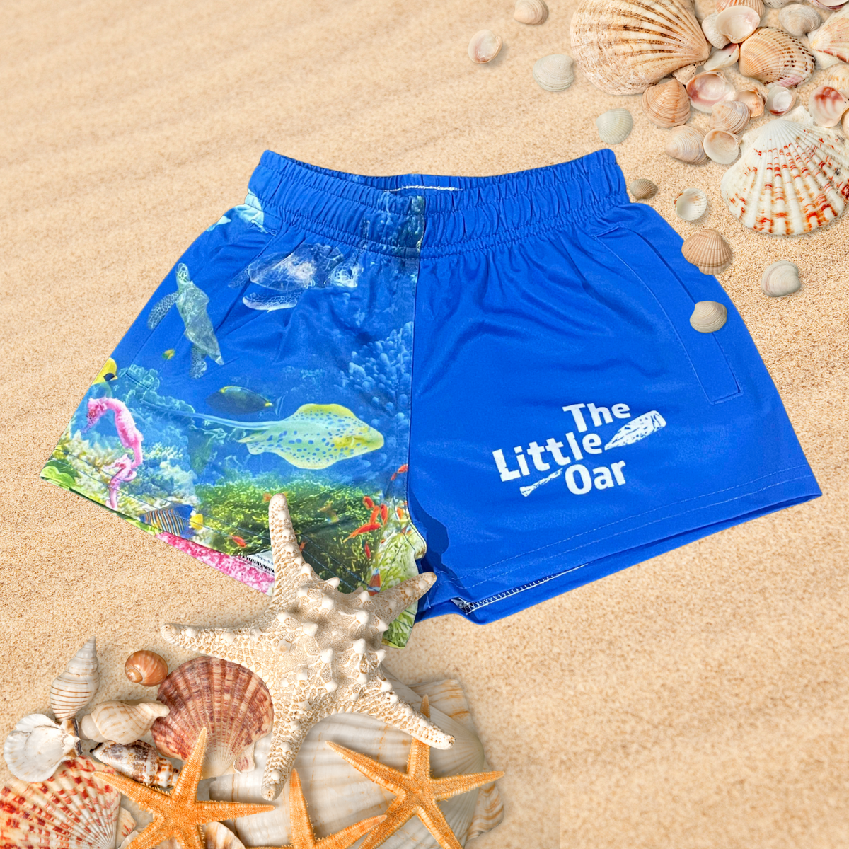 The Dirty Oars The Reef Kids Shorts on a background of sand and shells.
