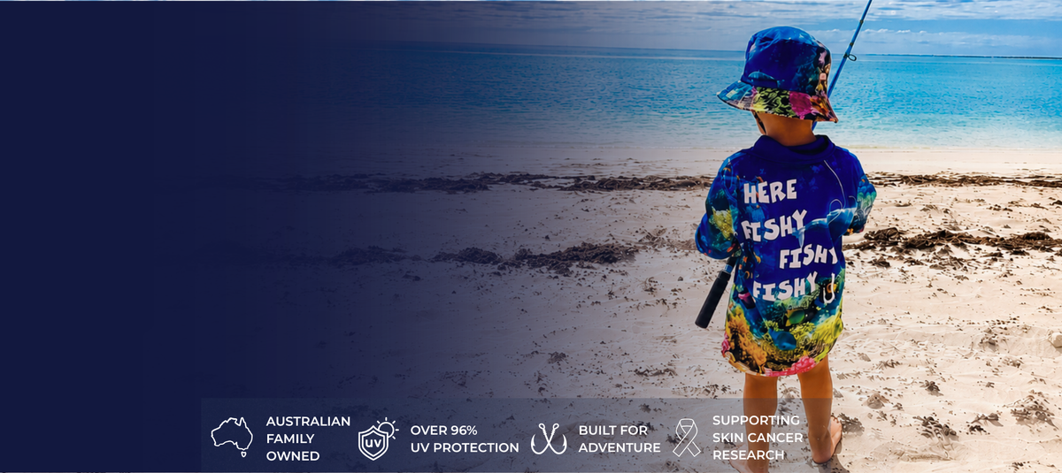 A child stands on a sandy beach wearing a colorful blue and yellow fishing-themed shirt with the text 'HERE FISHY FISHY FISHY!' and a matching hat, holding a fishing rod. The left side of the image has a dark blue gradient overlay with white text reading 'AUSTRALIAN FAMILY OWNED', 'OVER 96% UV PROTECTION', 'BUILT FOR ADVENTURE', and 'SUPPORTING SMALL FISHING FISHING'.