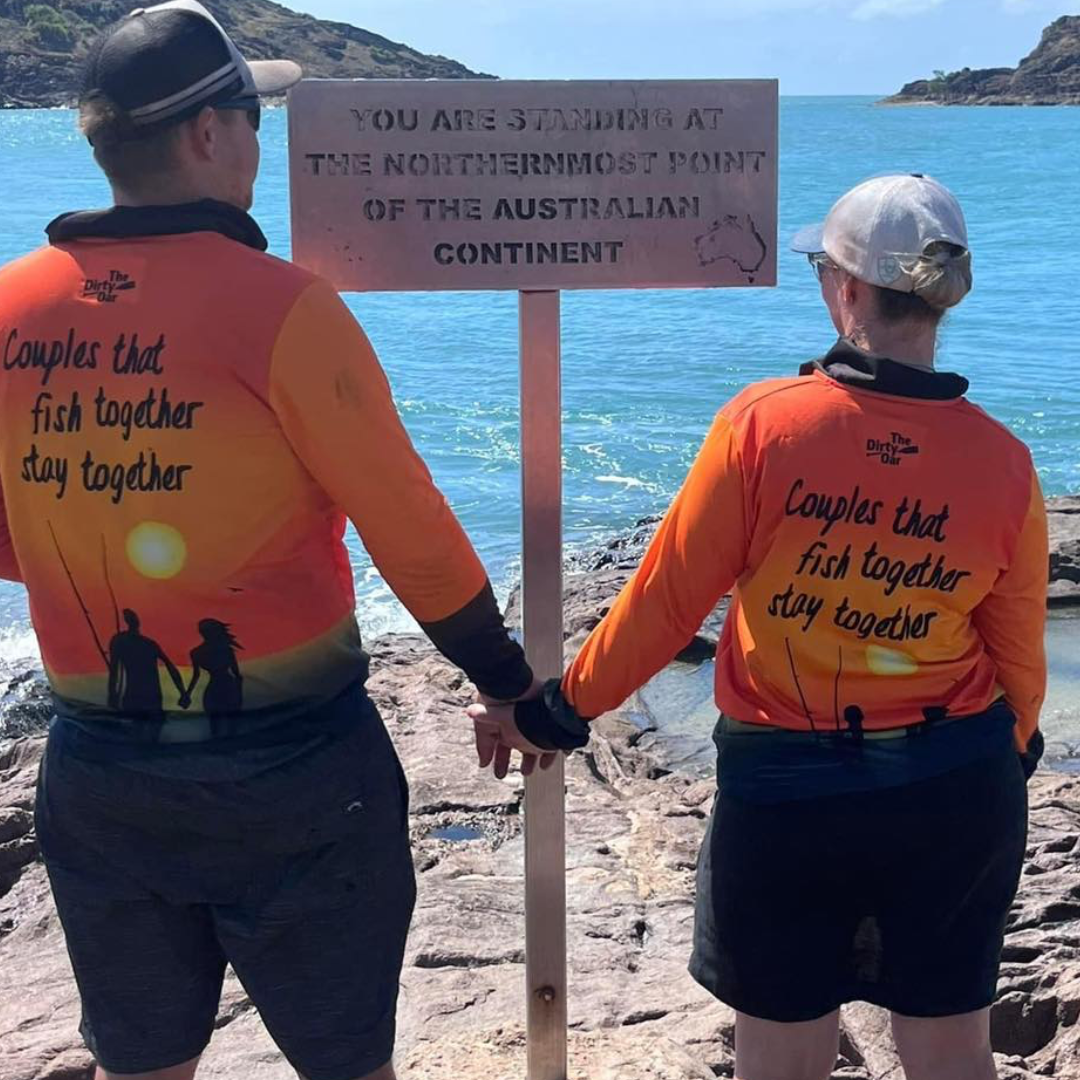 A couple wearing The Dirty Oars Couples Fishing Shirt standing in front of a sign at Tip of Australia Cape York.