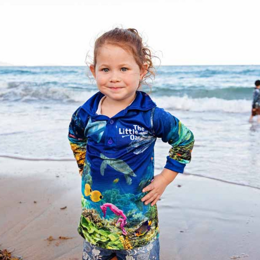 A child standing on a beach wearing The Dirty Oars The Reef Kids Fishing Shirt. 