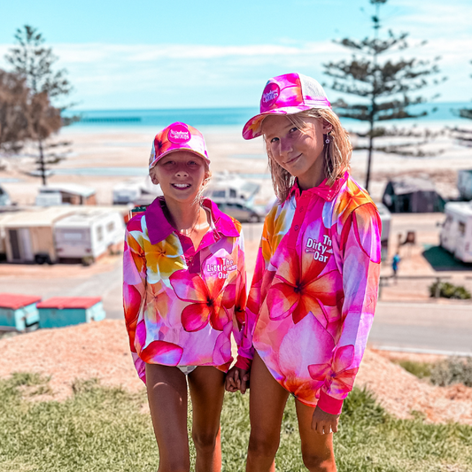 Two girls wearing The Dirty Oars Frangipani Kids Fishing Shirt and Frangipani Ponytail trucker caps, standing near a beach.
