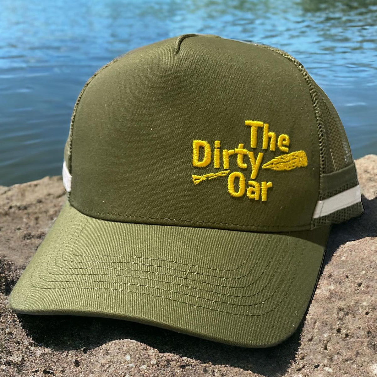 The Dirty Oar Camo Green Trucker Cap on rock near water.