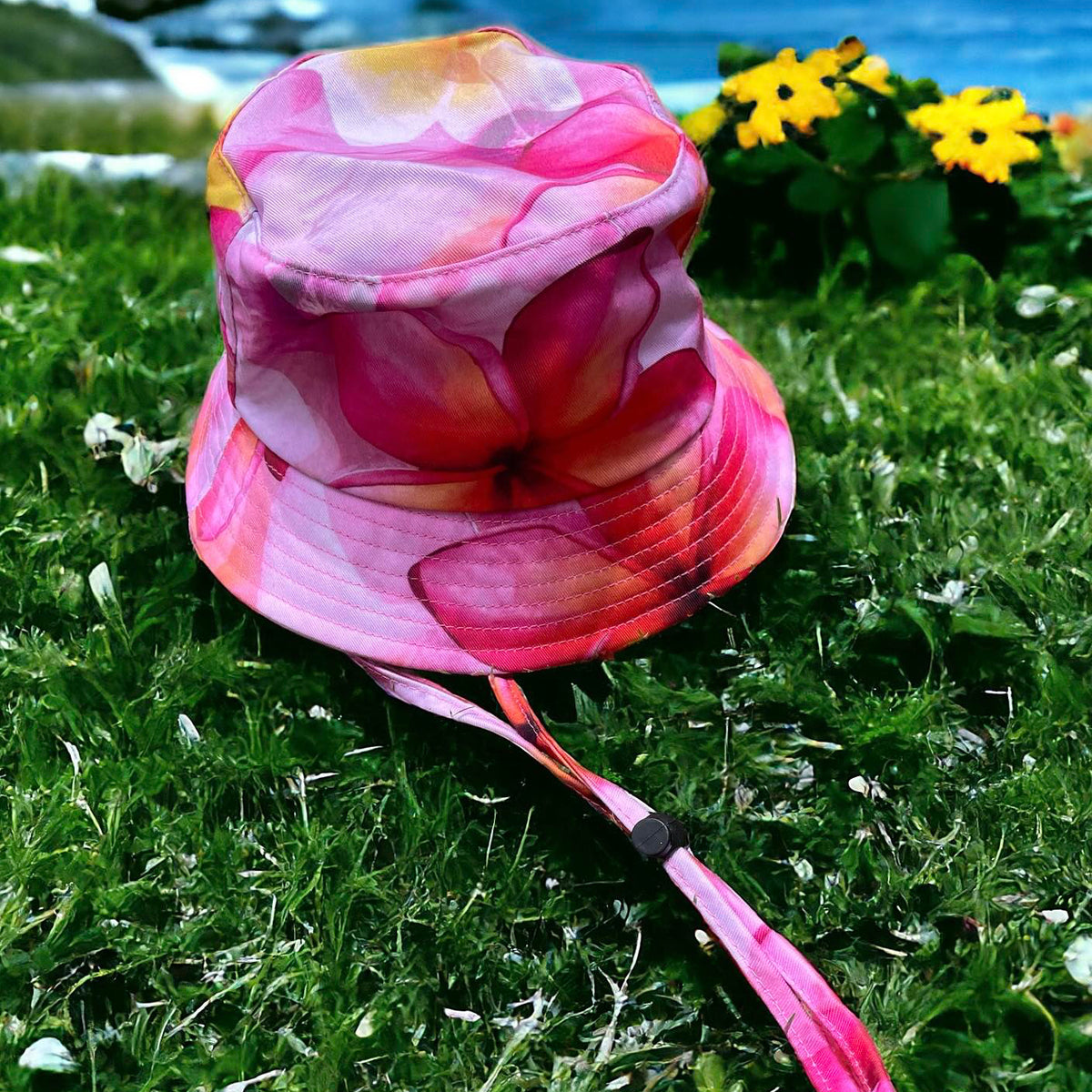 Frangipani Kids Bucket Hat is laid on green grass with a body of water and yellow flowers in the background.