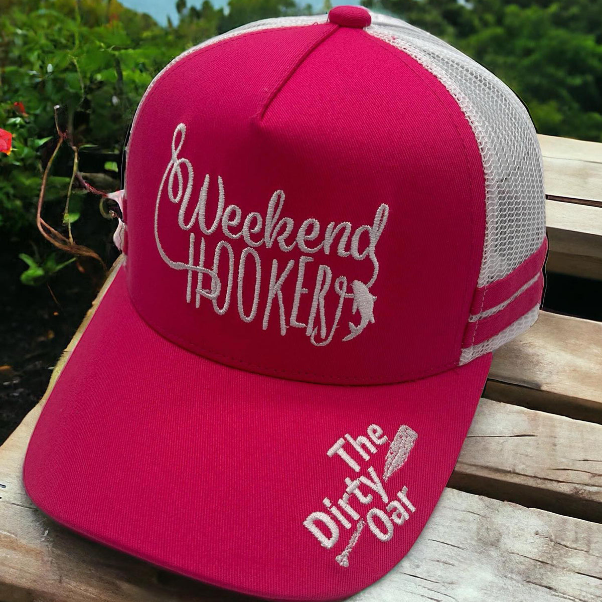 Weekend Hooker Ponytail Trucker Cap Pink The hat is displayed on a wooden surface with a scenic background.